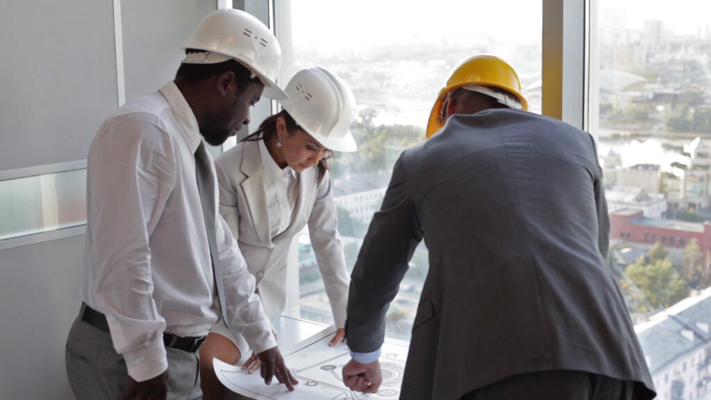 3 professionas in business attire, one in a suit with a yellow hard hat, the other two in white hard hats, poring over an architectural drawing or construction schematic