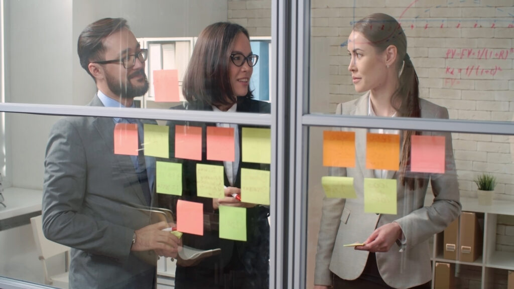 3 professionals in business attire stand in a glass room, having a meeting of sorts, with various coloured post-it notes stuck against the glass wall closet to the foreground