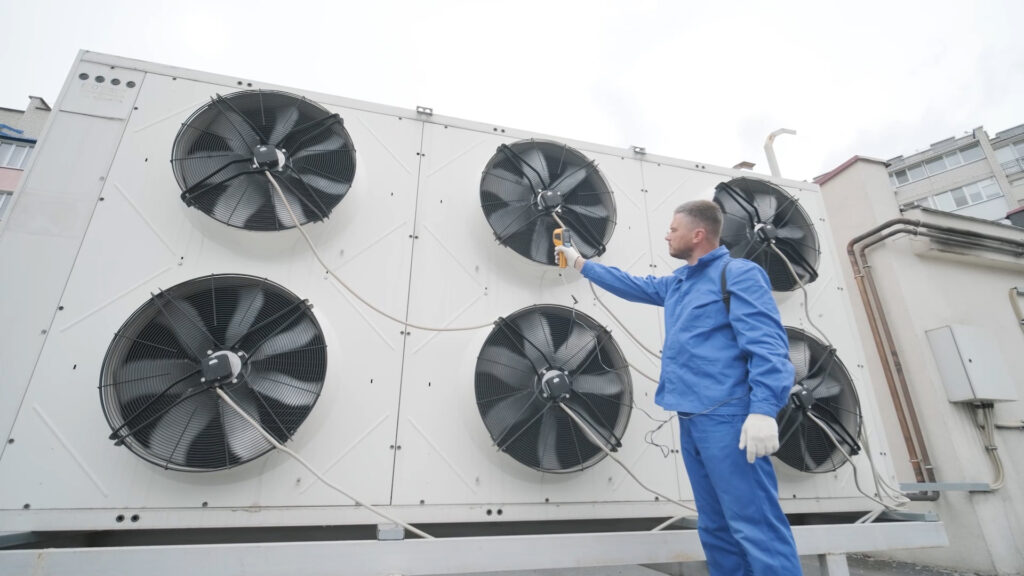 stock photo of large commercial HVAC units with large turbine fans, a technician in a blue uniform with white gloves points a laser thermometer at one of the fans reading its temperature