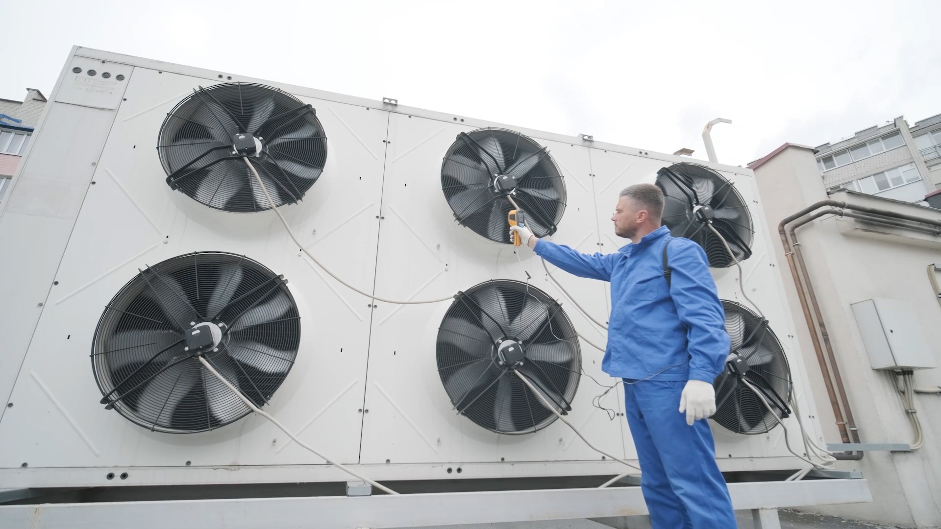 stock photo of large commercial HVAC units with large turbine fans, a technician in a blue uniform with white gloves points a laser thermometer at one of the fans reading its temperature