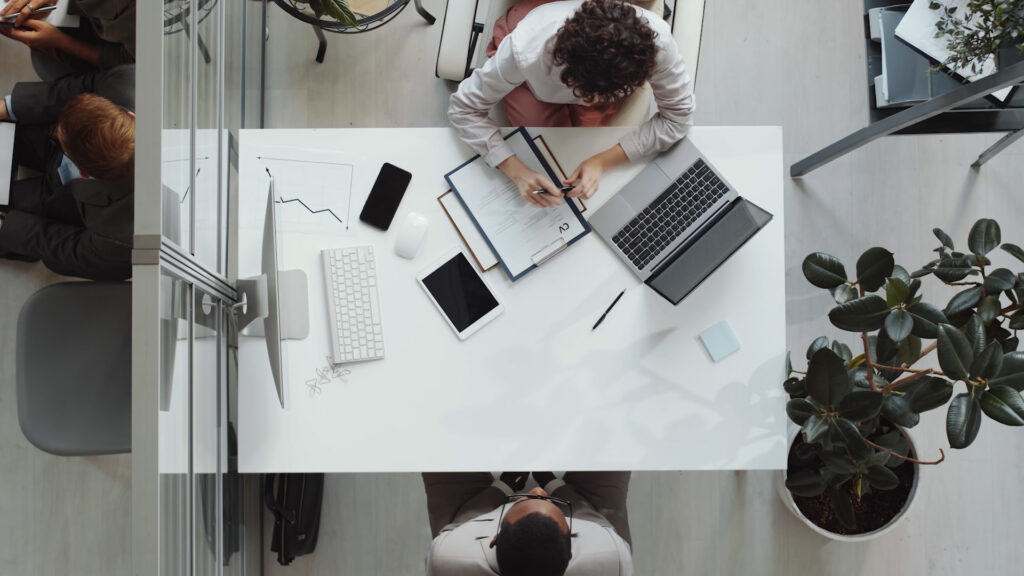 stock photo shot from above of two professionals seemingly in a job interview, a paper marked CV in front of one person (interviewer) and the interviewee sitting across the desk, which has two computers, a cell phone and a tablet