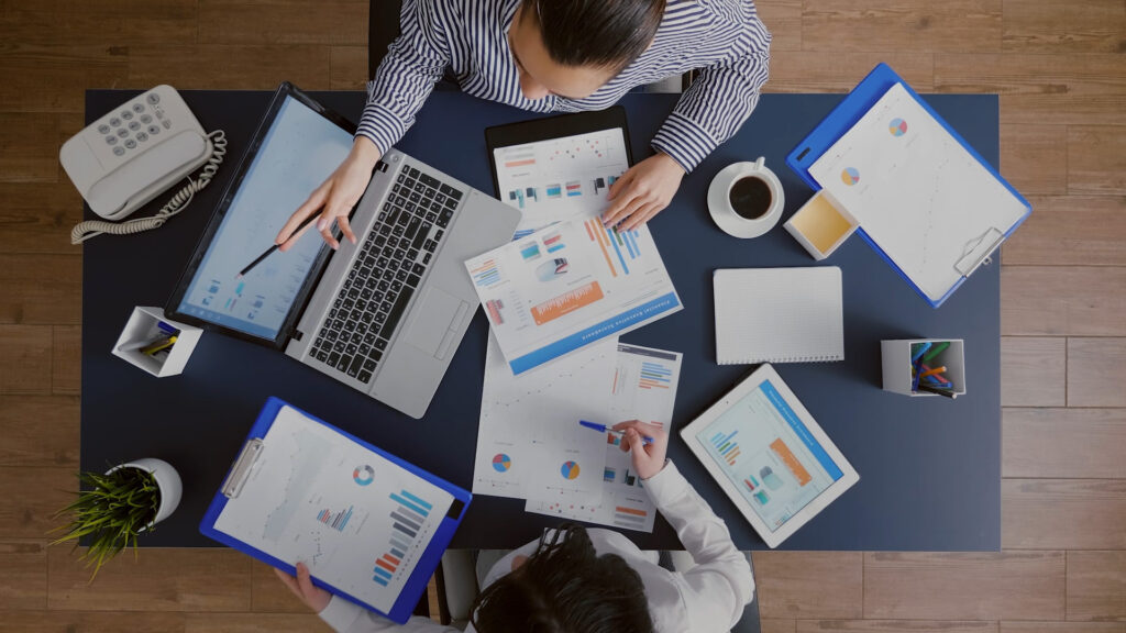 stock photo taken from overhead, of two professionals, poring over charts and graphs and data points, with one person pointing a their open laptop screen indicating the other should look at it