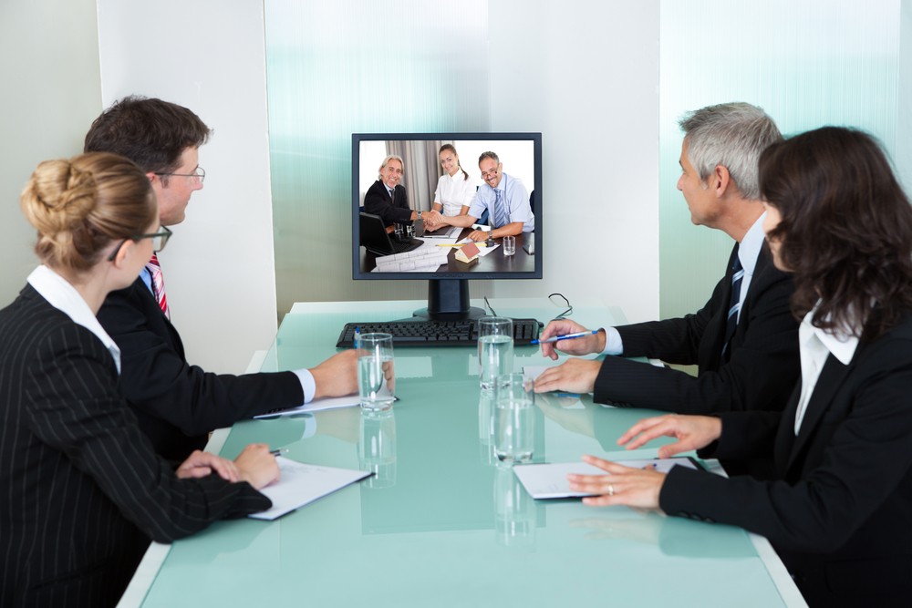 four professionals in business attire seated at a glass table together, taking in an online presentation on a monitor