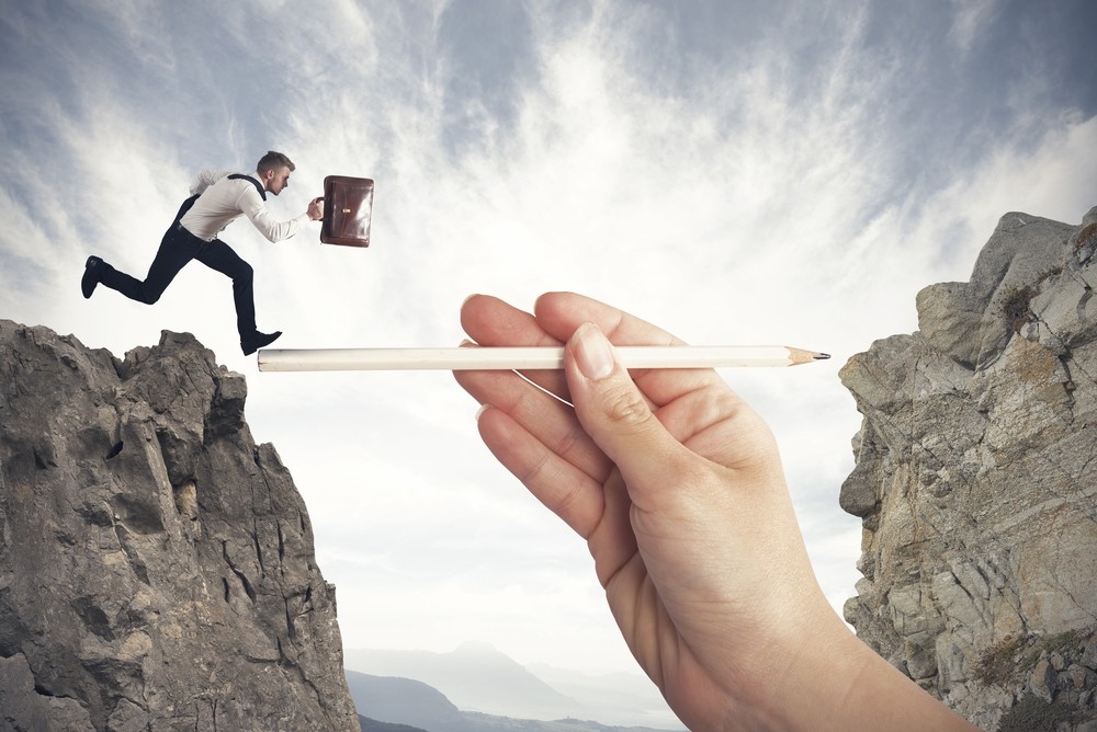 man in business attire holding a briefcase running across a chasm on a bridge made up of a pencil being held by a larger than life hand