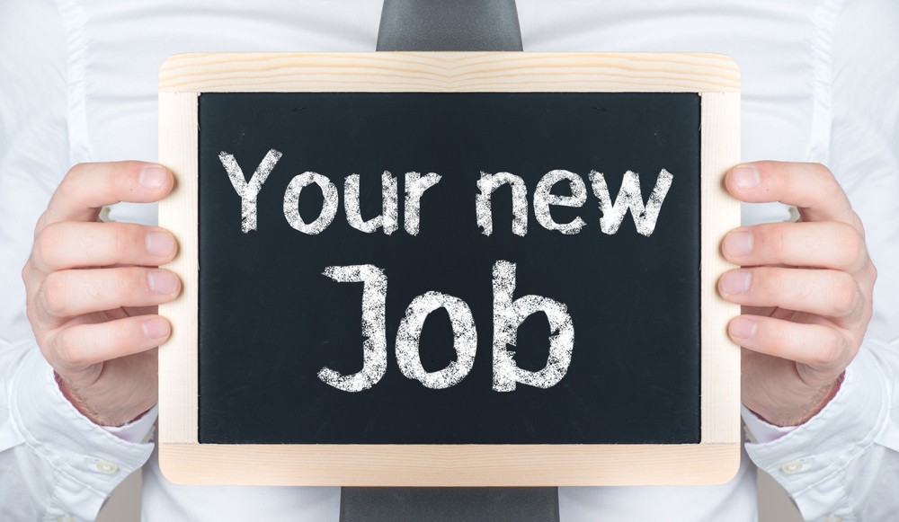 person in white shirt and black tie holding a chalkboard with both hands, upon which is inscribed "Your new Job"