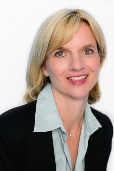 Photo of Judy Geerts, dark suit jacket, light turqouise dress shirt unbuttoned at the top, shoulder length blonde hair, smiling